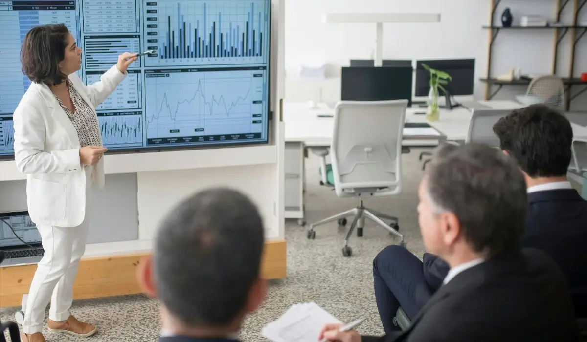 Business consultant presenting data-driven strategies to Fortune 500 executives in a corporate meeting room. Charts and graphs displayed on a large screen.