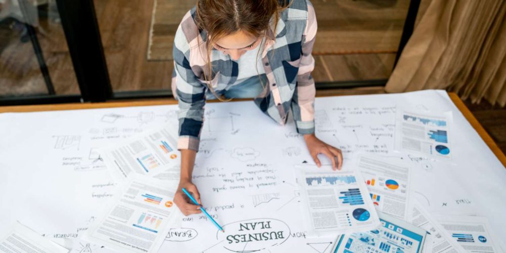 woman at a large table with multiple papers spread out on a table putting together a business plan
