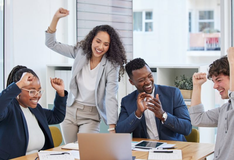 four people sitting at a desk smiling with their hands in the air after achieving a goal